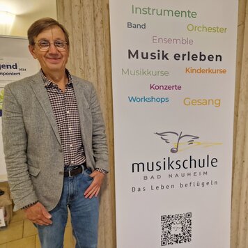 Ein freundlich lächelnder Mann mit Brille steht neben einem Werbebanner der Musikschule Bad Nauheim. | © BNST GmbH