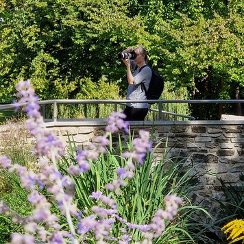 Blonde Frau mit Rucksack steht auf einer Steinbrücke in einem Park und fotografiert mit einer Spiegelreflexkamera.  | © BNST GmbH