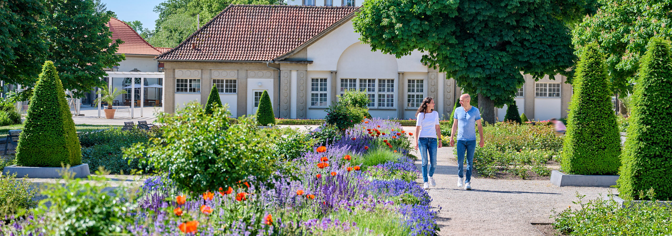 Eine Frau und ein Mann gehen in einem Garten spazieren, in dem Klatschmohn, Lavendel, Rosen und Buchsbäume blühen. | © Florian Trykowski 